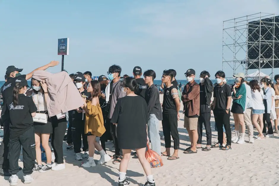 A large group of people wearing masks stand in line at a beach event entrance under clear skies.