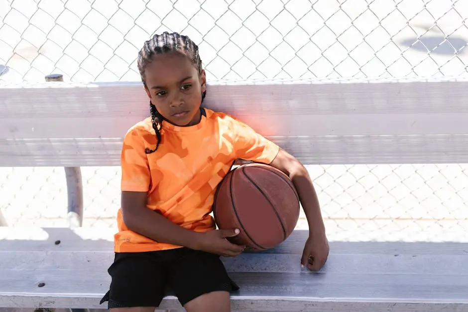 A young boy sitting with a basketball on an outdoor bench in casual attire.