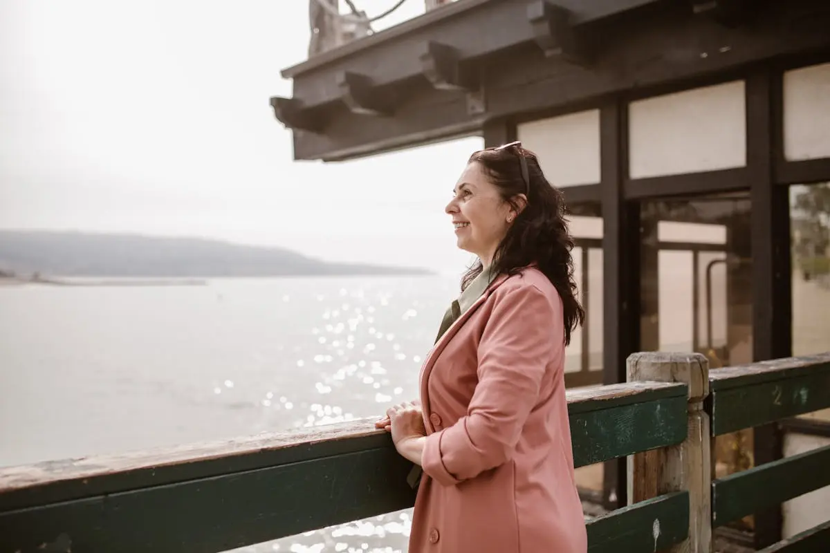 Smiling woman in pink coat enjoying a peaceful moment by the water’s edge.