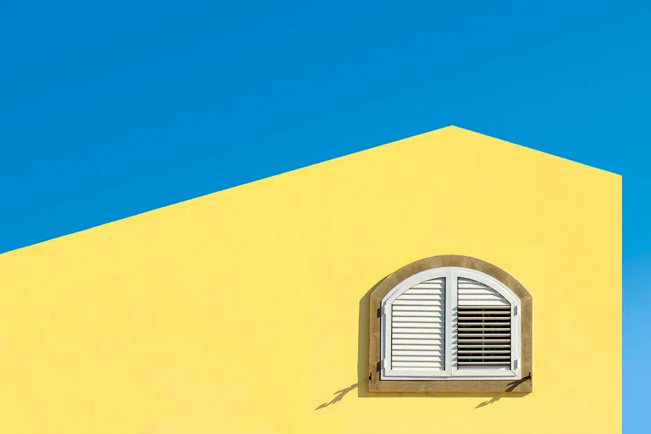 Bright yellow wall with an arched white shuttered window against a clear blue sky.