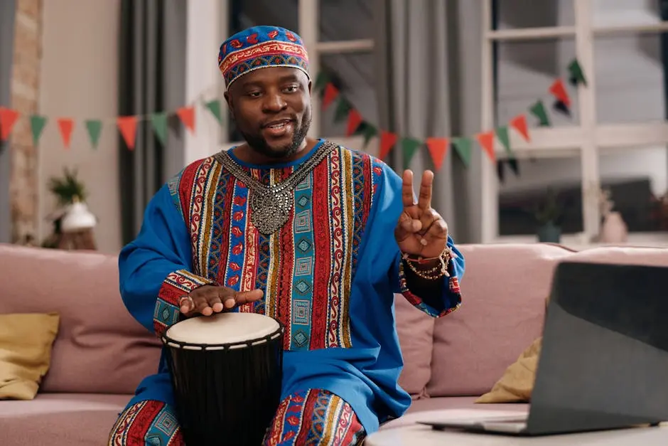 African man wearing traditional attire playing djembe drum and celebrating Kwanzaa indoors.