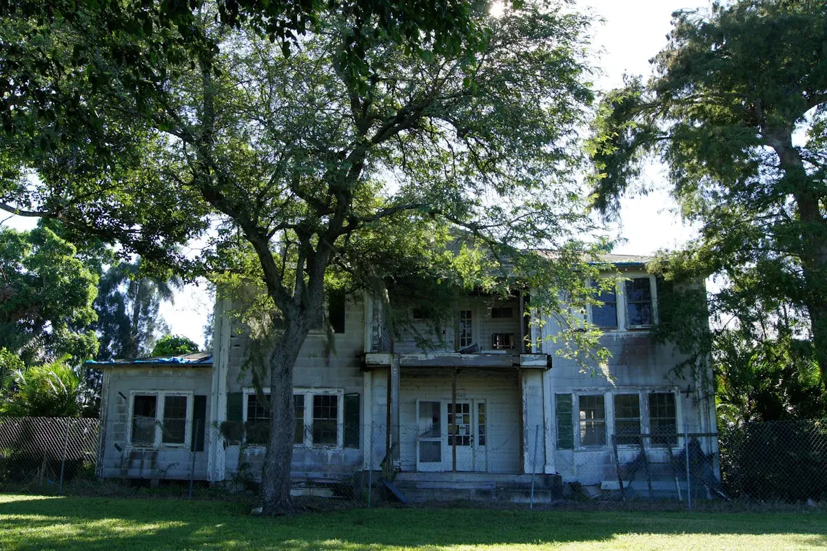 Dilapidated two-story house amidst lush greenery in the afternoon sunlight.