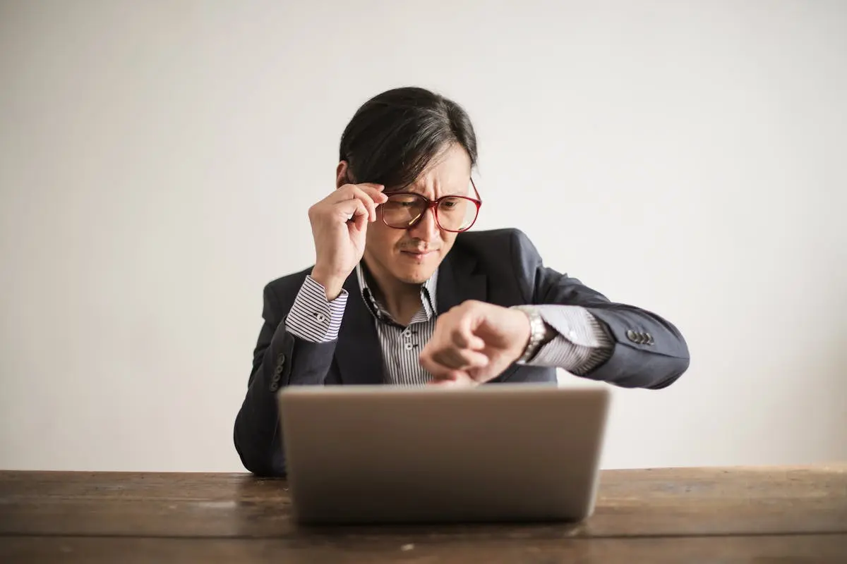 Young frowning man in suit and glasses looking at wristwatch while waiting for appointment sitting at desk with laptop