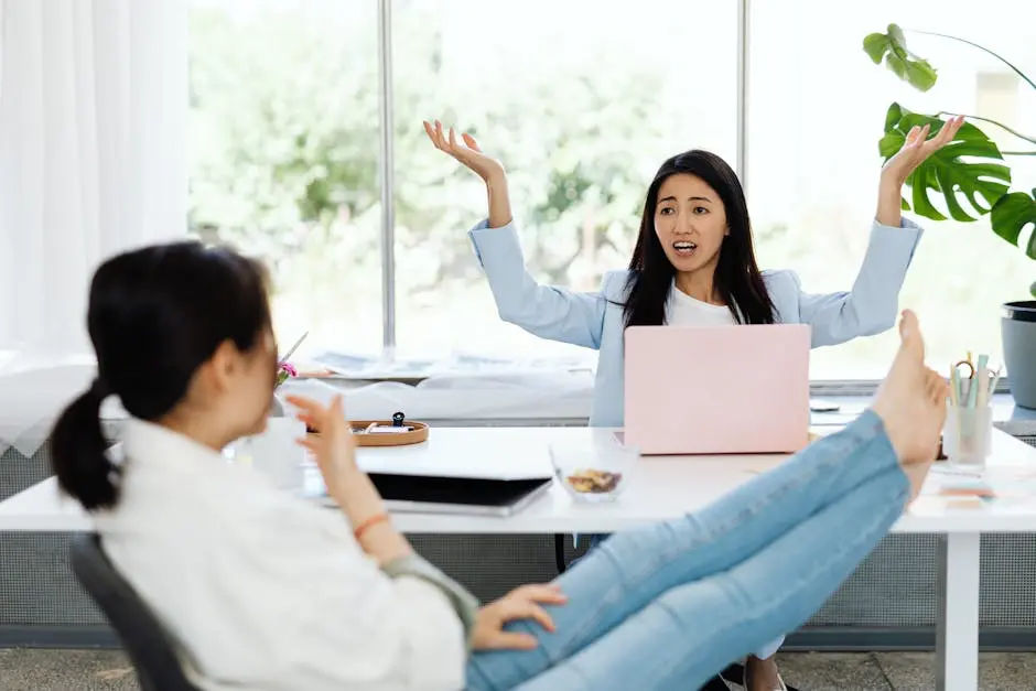 Two women engage in an animated discussion in a modern office setting, surrounded by laptops and relaxed atmosphere.
