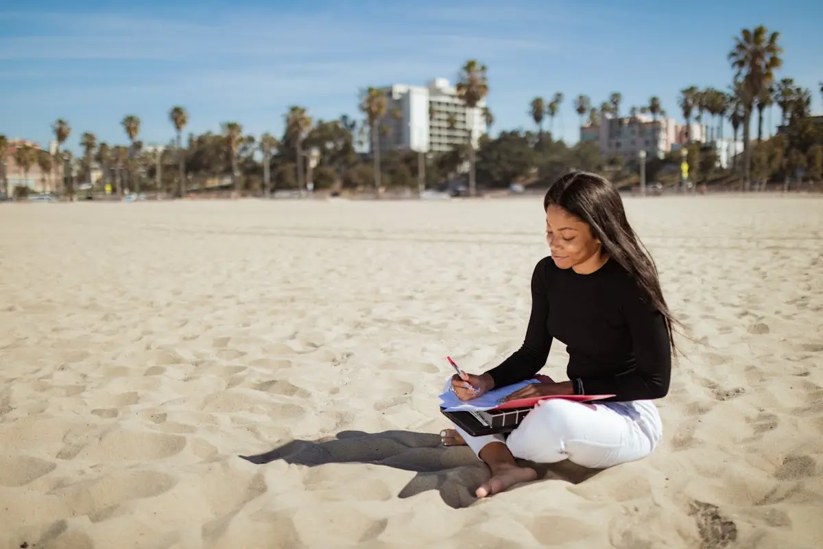 Woman writing on documents with tablet on a sunny beach, showcasing remote work possibilities.