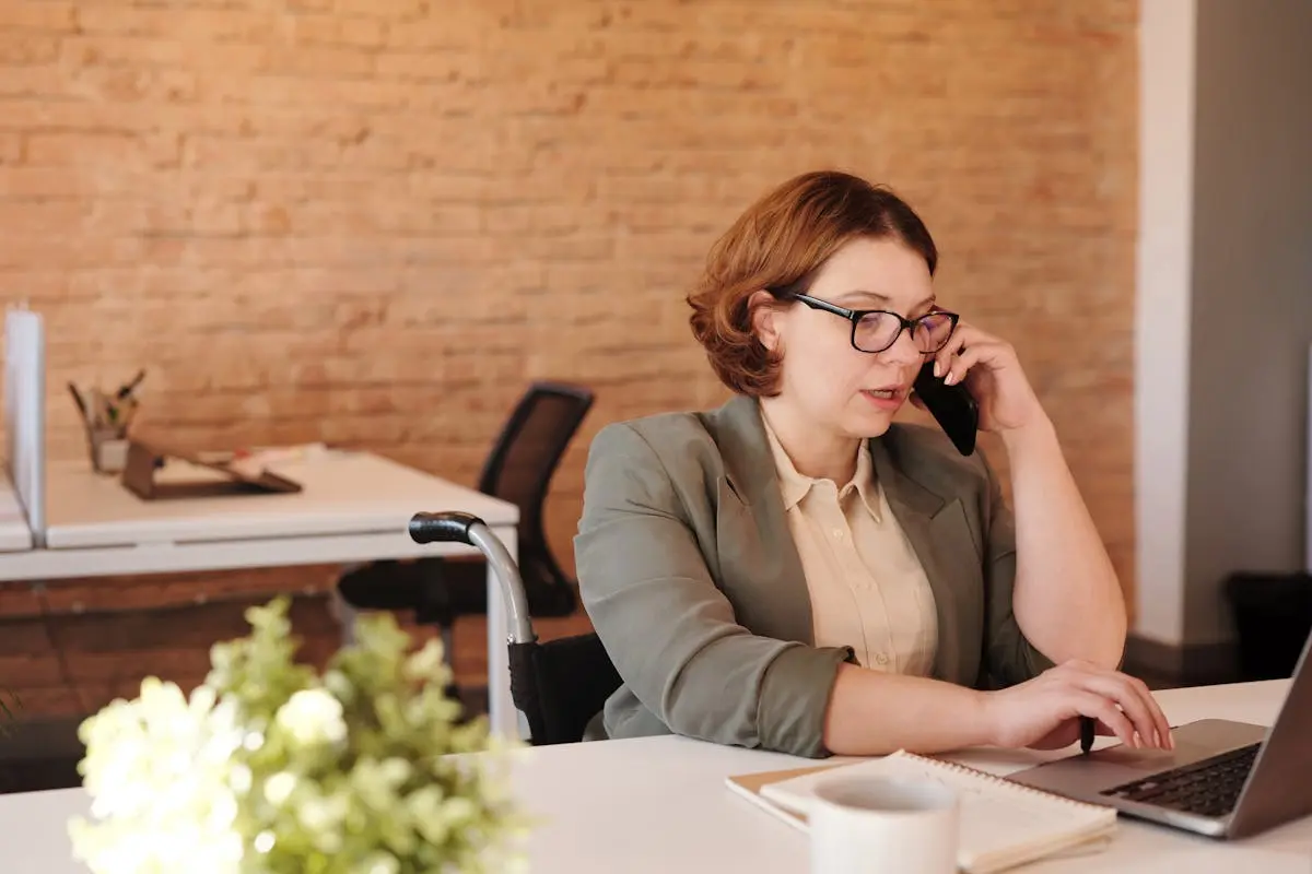 Professional woman in wheelchair working remotely on laptop and phone in a modern office.