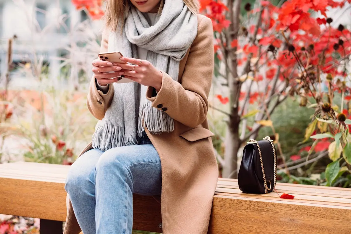 A woman in a coat and scarf texting on her phone while sitting on a bench in a park.