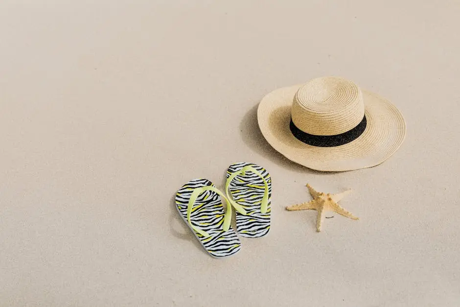Flat lay of beach accessories including a straw hat, zebra flip flops, and a starfish on sandy surface.