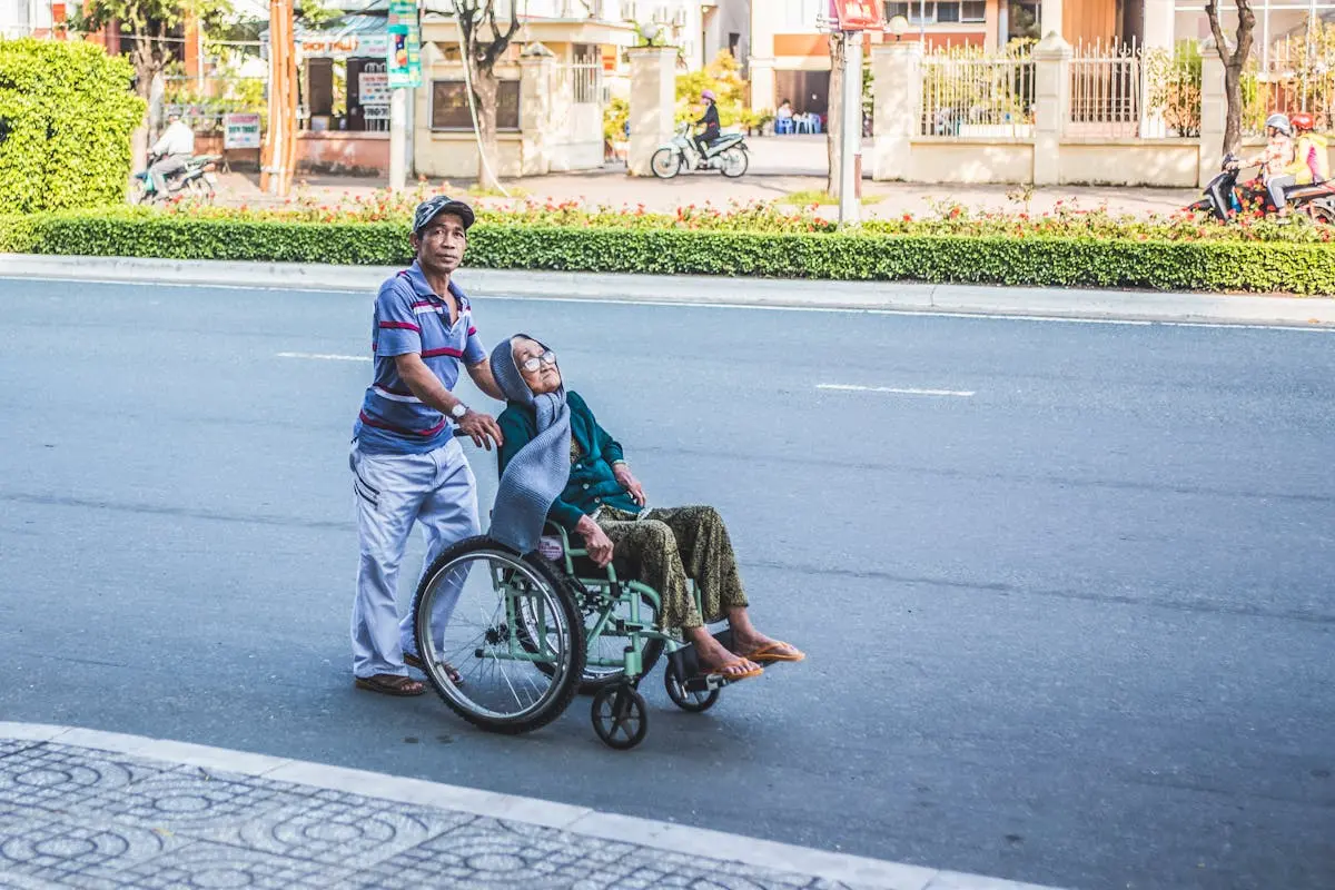 A caring man aids an elderly woman in a wheelchair across a sunny urban street in Cần Thơ, Vietnam.