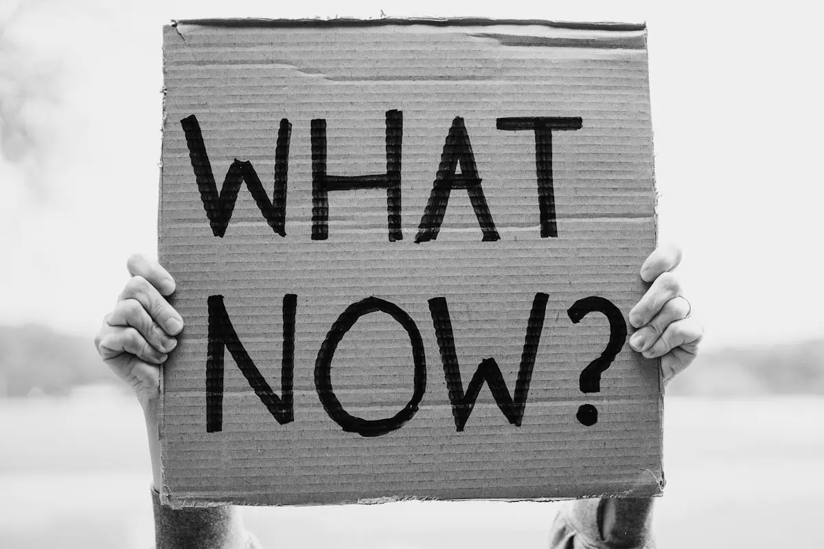A close-up of hands holding a cardboard sign with the text ‘What Now?’