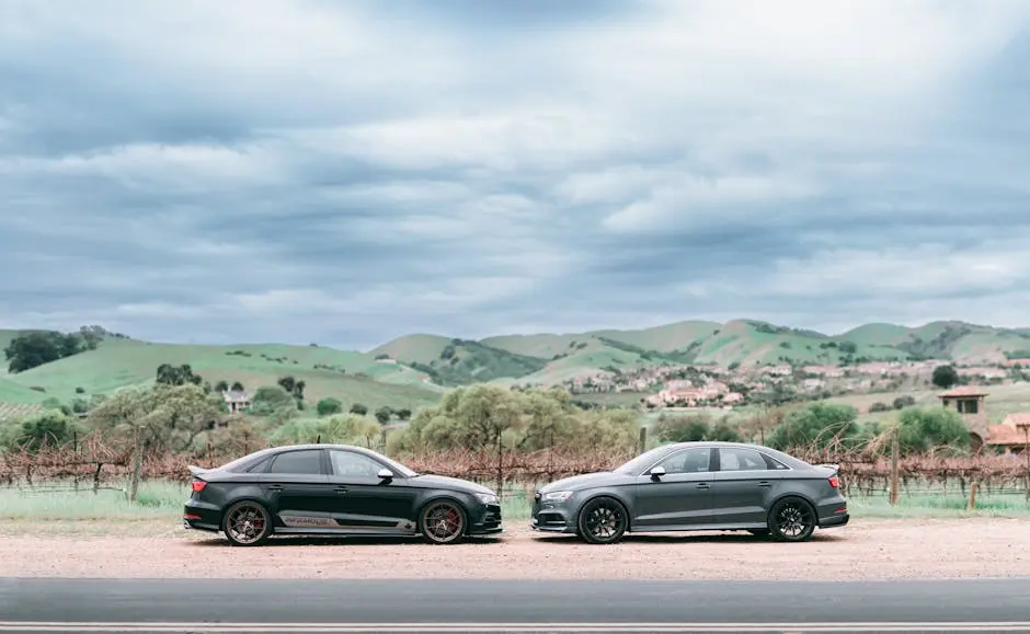 Two Audi cars parked on a scenic road with rolling hills and cloudy sky in the background.