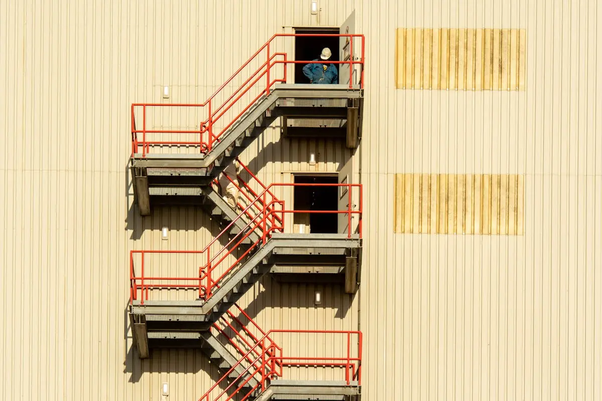 Worker in hardhat standing on fire escape stairs of industrial building with beige walls.