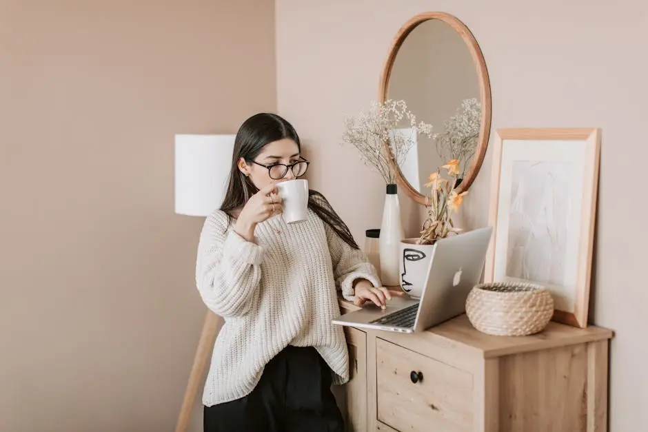 A young woman enjoys coffee while working on her laptop in a cozy home environment.