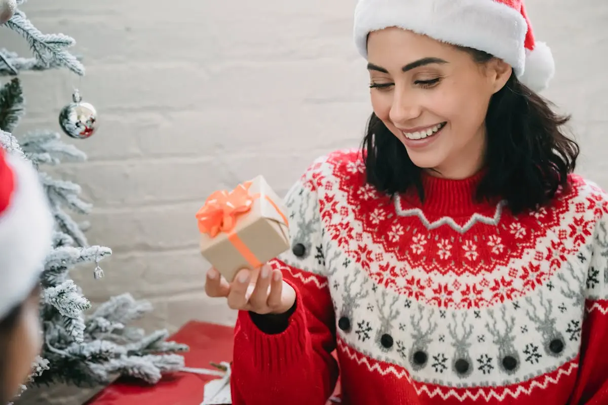 Joyful woman in Santa hat unwrapping a Christmas gift by the decorated tree.