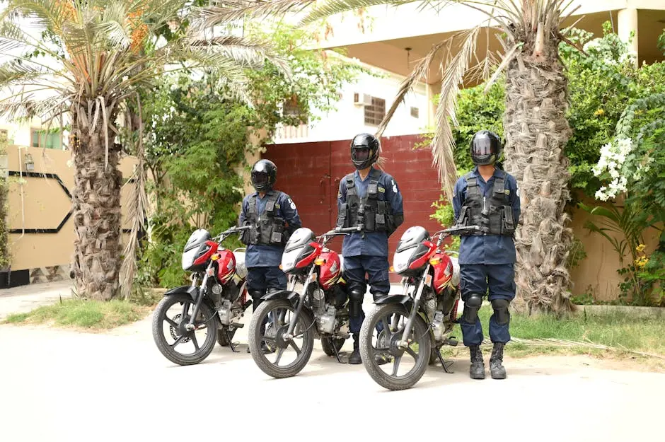 Three security guards in uniform standing by motorbikes outdoors in a tropical setting.
