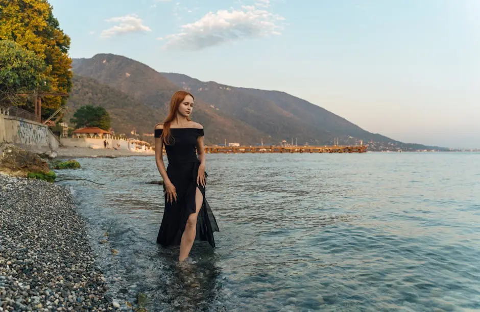 Elegant woman in a black dress strolls along the beach in Гагра with a mountain backdrop during sunset.
