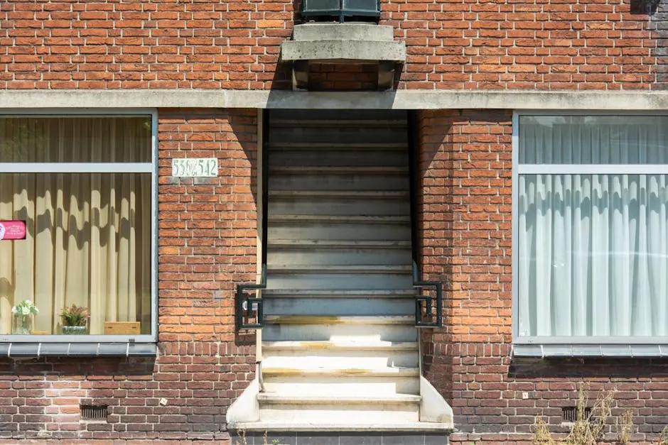 A close-up of a brick residential building facade featuring stairs and large windows with curtains.