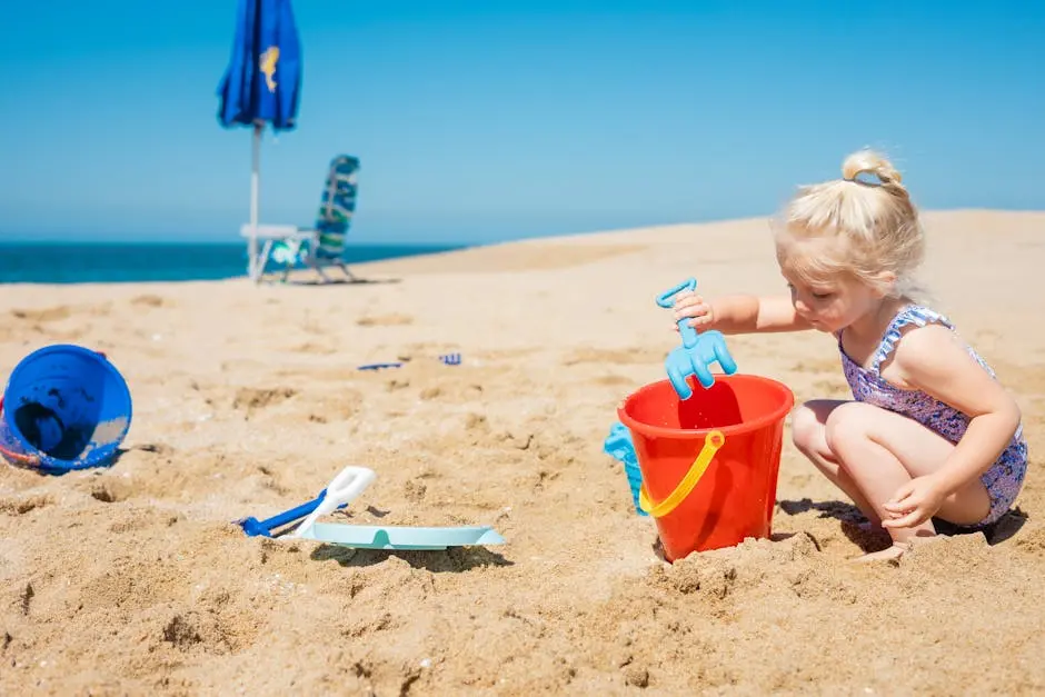 Young girl playing with red bucket and spade on a sunny beach day, enjoying summer fun.