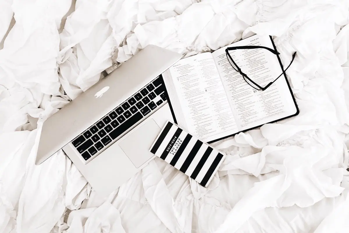 A flat lay of a laptop, open book, and reading glasses on a white bed. Perfect for productivity and relaxation themes.