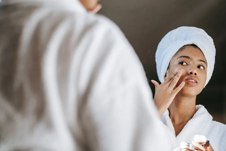 A young woman applies skincare cream while wearing a bathrobe in front of a mirror.