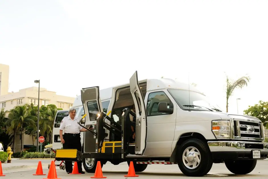A driver standing by a wheelchair accessible van with open doors and a ramp, surrounded by cones.