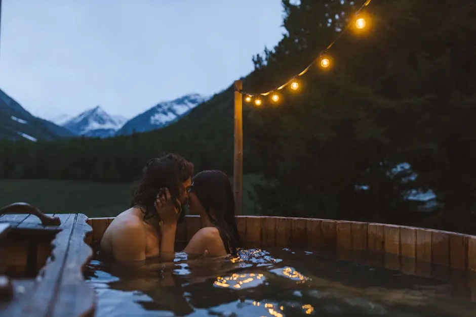 A couple shares a romantic kiss in a cozy outdoor hot tub surrounded by mountains at twilight.