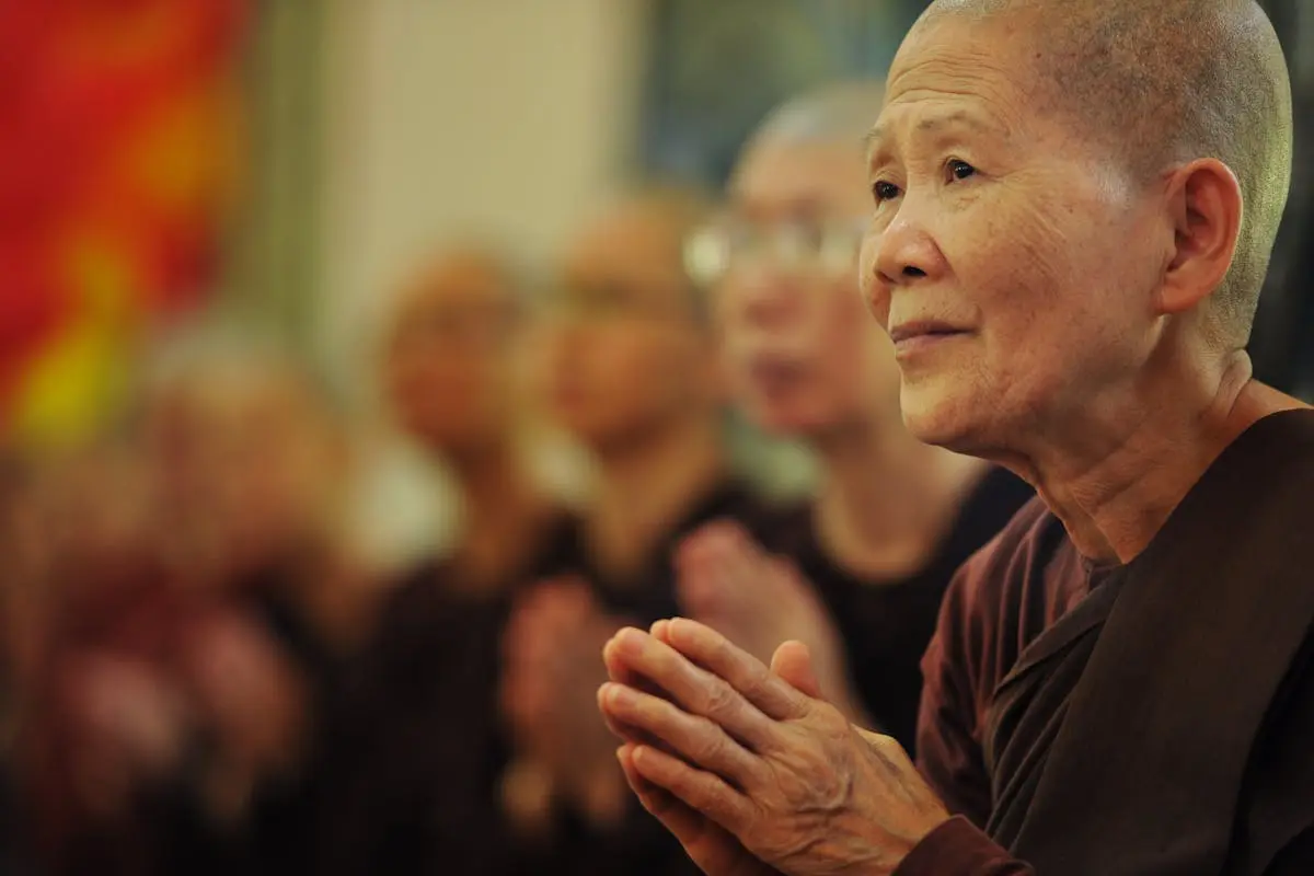 Senior Buddhist woman in prayer, surrounded by fellow practitioners, inside a temple.
