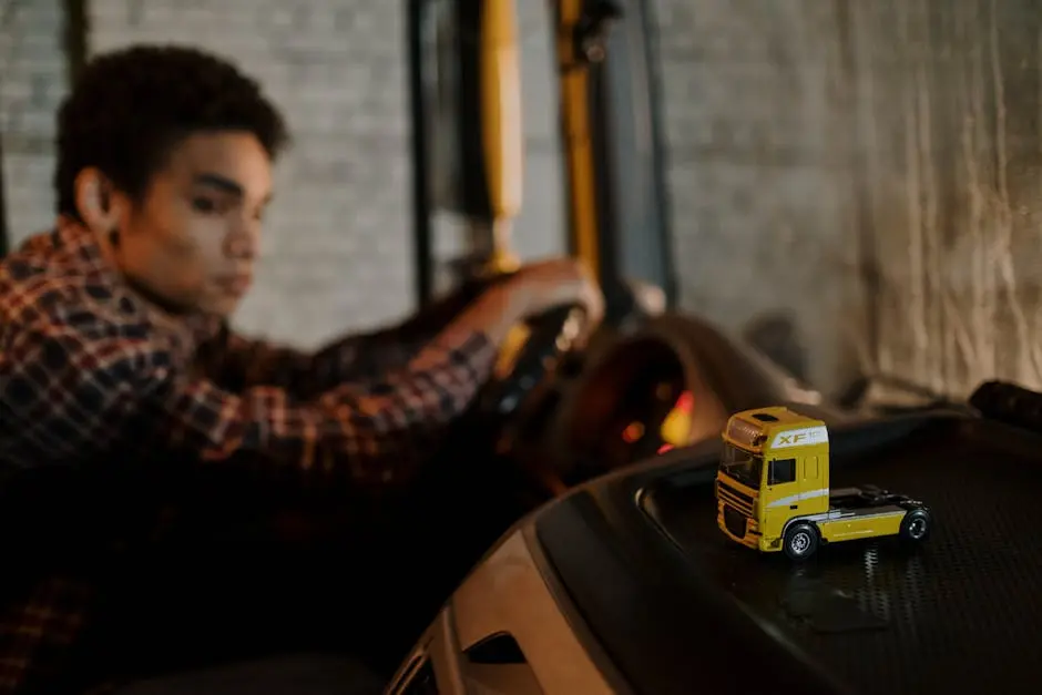 A driver in a truck cab with a focus on a miniature yellow truck toy, creating a blurred background.
