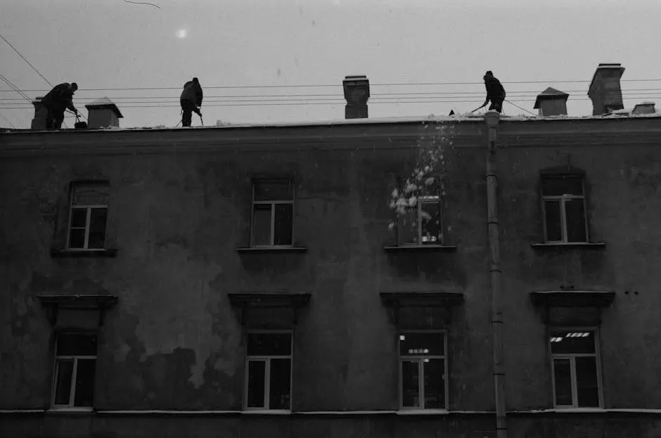 Silhouetted workers clear snow from a building rooftop during winter. Black and white photograph.
