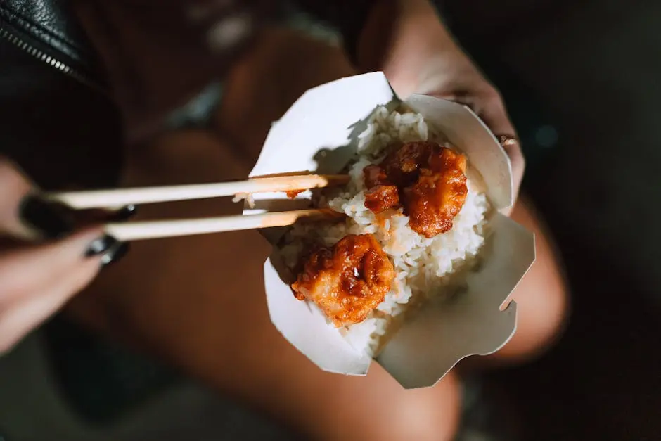 Fried shrimp with rice in a takeout box, held with chopsticks, exuding Asian culinary appeal.