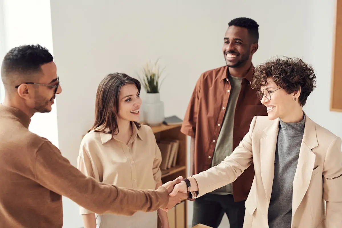Four colleagues smiling and small business consulting and shaking hands in a bright office setting.